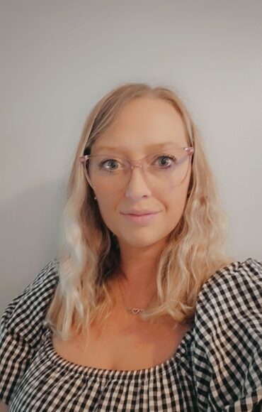 A woman wearing glasses and a black and white checked top, smiling confidently against a neutral background.