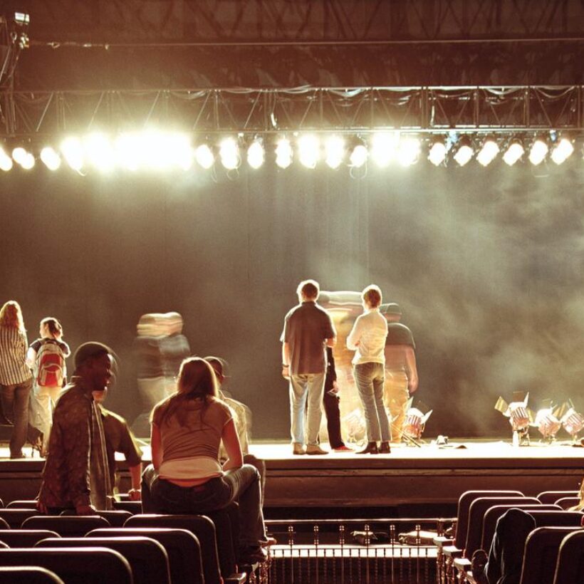 A brightly lit stage with performers rehearsing, while a crowd observes the performance