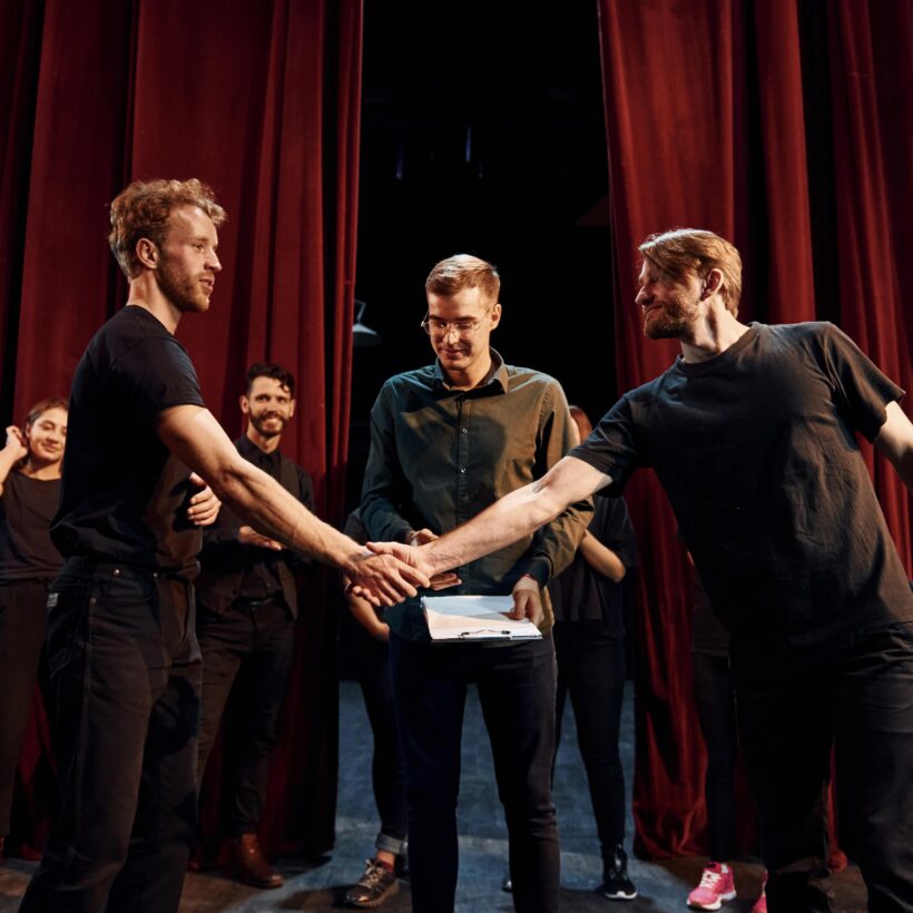 On stage, two men shake hands in front of another man against a curtain backdrop.