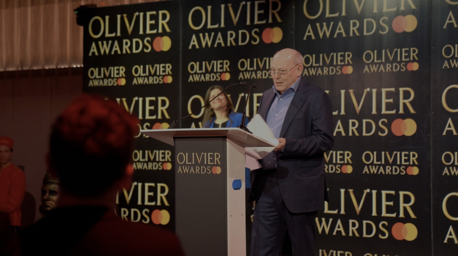 A man with glasses dressed in a suit standing at a podium delivering and acceptance speech